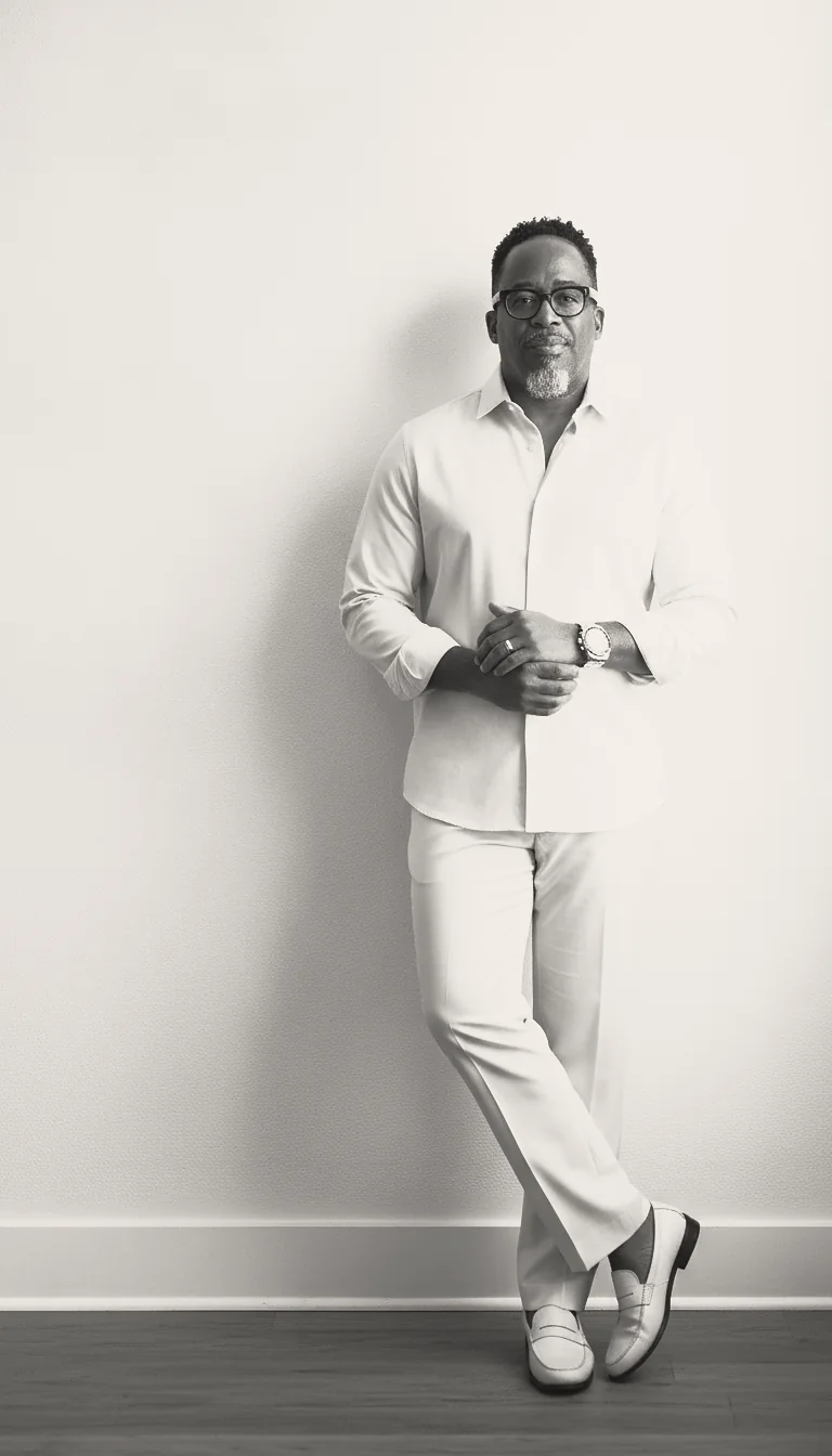 Black and white full-length portrait of Kenneth leaning against a white wall, adjusting his watch